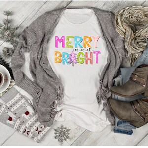 K&ouml;nnte beinhalten: Ein wei&szlig;es T-Shirt mit den Worten "MERRY and BRIGHT" in bunten Blockbuchstaben und einer Weihnachtsbaumgrafik. Das Shirt wird mit einer grauen Strickjacke, Jeans, Stiefeln und einem Schal kombiniert, was auf ein festliches, l&auml;ssiges Outfit hindeutet.