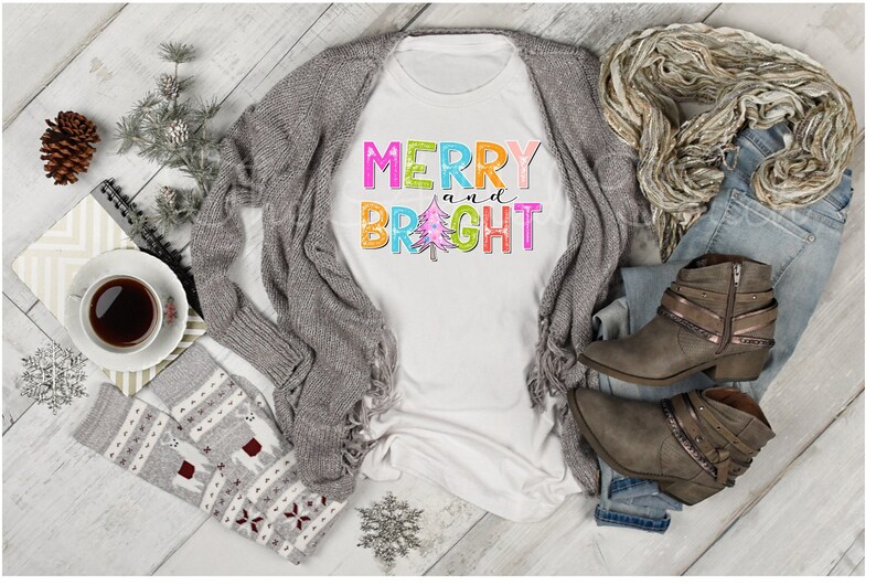 K&ouml;nnte beinhalten: Ein wei&szlig;es T-Shirt mit den Worten "MERRY and BRIGHT" in bunten Blockbuchstaben und einer Weihnachtsbaumgrafik. Das Shirt wird mit einer grauen Strickjacke, Jeans, Stiefeln und einem Schal kombiniert, was auf ein festliches, l&auml;ssiges Outfit hindeutet.