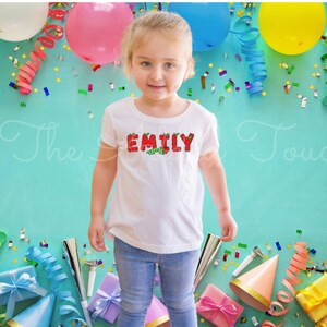 May include: A young girl wearing a white t-shirt with the name "EMILY" spelled out in red letters with green leaves. The letters are decorated with a caterpillar design. The girl is surrounded by colorful party decorations, including balloons, streamers, and confetti.
