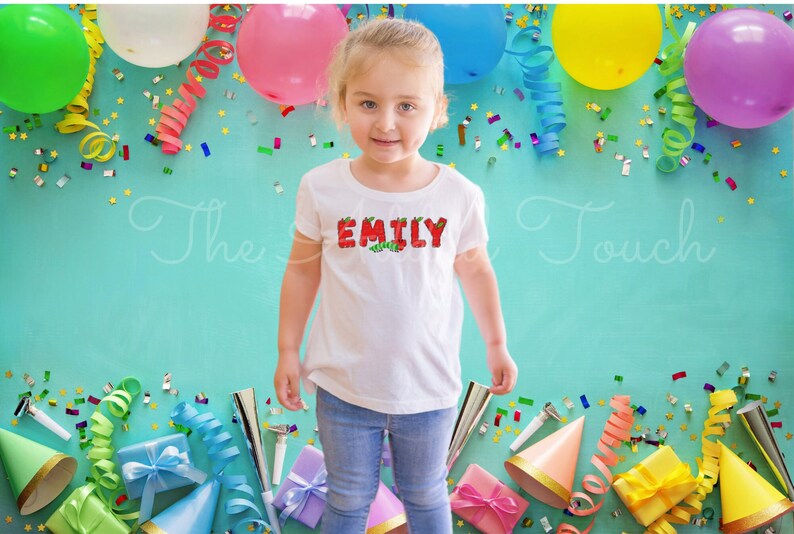 May include: A young girl wearing a white t-shirt with the name "EMILY" spelled out in red letters with green leaves. The letters are decorated with a caterpillar design. The girl is surrounded by colorful party decorations, including balloons, streamers, and confetti.