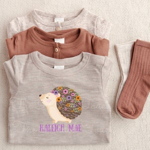 May include: A stack of baby clothes including a light gray onesie, a brown onesie, and a light gray t-shirt with a hedgehog graphic. The hedgehog has a floral crown. Two pairs of socks in light gray and brown are also included.
