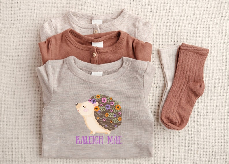 May include: A stack of baby clothes including a light gray onesie, a brown onesie, and a light gray t-shirt with a hedgehog graphic. The hedgehog has a floral crown. Two pairs of socks in light gray and brown are also included.