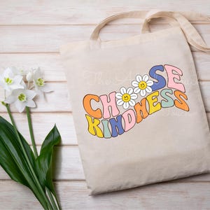 May include: A beige tote bag featuring the text "Choose Kindness" in a rainbow of colours and a daisy design. The bag has a long strap and is made of canvas.