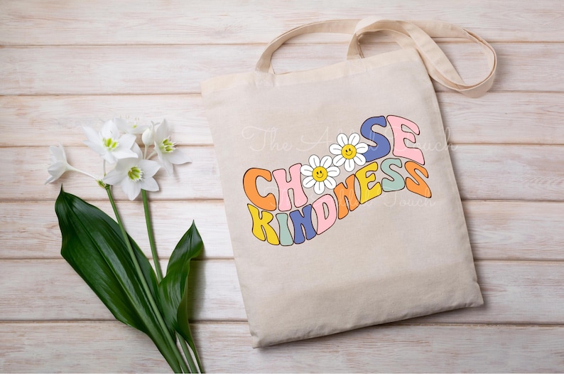 May include: A beige tote bag featuring the text "Choose Kindness" in a rainbow of colours and a daisy design. The bag has a long strap and is made of canvas.