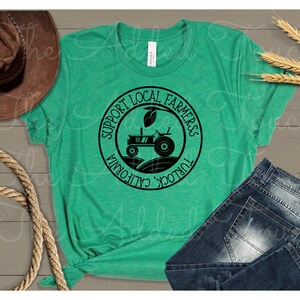 Pu&ograve; includere: Una maglietta verde con un grafico circolare nero che dice "Support Local Farmers Turlock, California". Il grafico presenta anche una silhouette nera di un trattore in un campo.