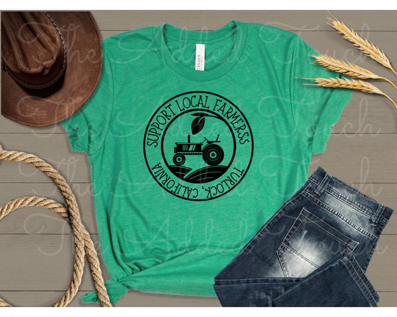 Pu&ograve; includere: Una maglietta verde con un grafico circolare nero che dice "Support Local Farmers Turlock, California". Il grafico presenta anche una silhouette nera di un trattore in un campo.
