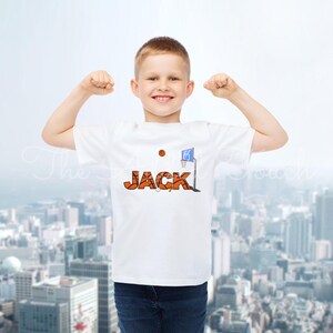 May include: A white t-shirt featuring the name "JACK" in orange basketball-themed lettering, a basketball, and a blue and white basketball hoop graphic. The boy is wearing blue jeans.