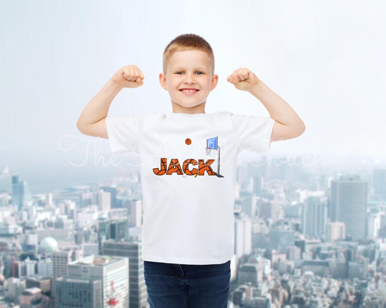 May include: A white t-shirt featuring the name "JACK" in orange basketball-themed lettering, a basketball, and a blue and white basketball hoop graphic. The boy is wearing blue jeans.