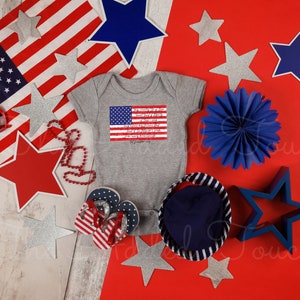 May include: A gray baby onesie with a red, white, and blue American flag and text on the front. The onesie is surrounded by red, white, and blue stars, a blue fan, and red and white heart-shaped sunglasses.