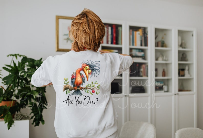 Pode incluir: Camisola branca com uma ilustra&ccedil;&atilde;o de um p&aacute;ssaro de desenho animado e o texto "Are You Okay?" impresso nas costas.