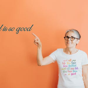 May include: A woman wearing a white t-shirt with the text "and all my life you have been faithful and all my life you have been so so good" in rainbow colors. The woman is pointing to the left and smiling.