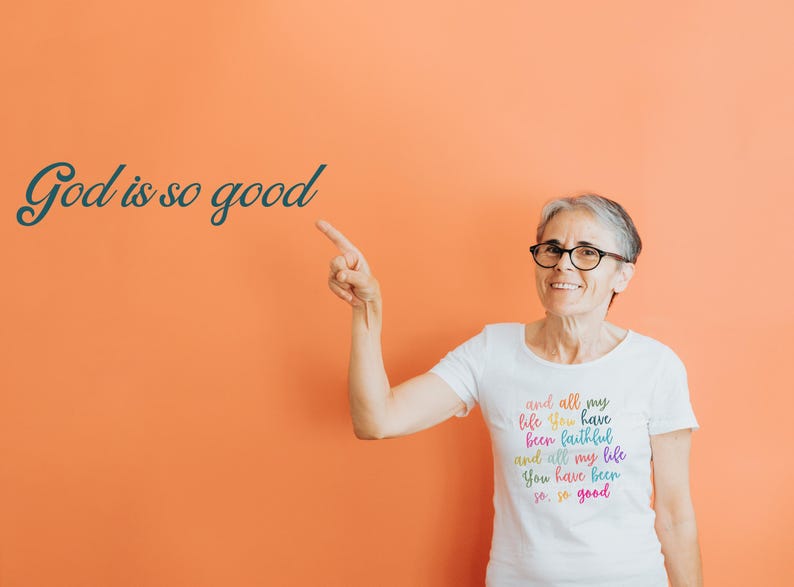 May include: A woman wearing a white t-shirt with the text "and all my life you have been faithful and all my life you have been so so good" in rainbow colors. The woman is pointing to the left and smiling.
