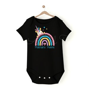 May include: Black baby onesie with a rainbow and a unicorn design. The rainbow is in shades of pink, yellow, blue, and green. The text "Makhaira Dianne" is printed below the rainbow.