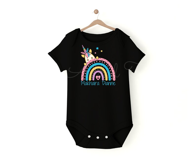 May include: Black baby onesie with a rainbow and a unicorn design. The rainbow is in shades of pink, yellow, blue, and green. The text "Makhaira Dianne" is printed below the rainbow.