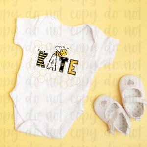 May include: White baby onesie with a bee and honeycomb design. The onesie has the name "KATE" spelled out in yellow and black letters. Two white baby shoes are on a yellow background.