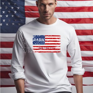 May include: White long-sleeve shirt with a distressed American flag design. The flag has the word "DADA" in blue lettering and the names "Charlie", "Claire", and "Cruz" in smaller blue lettering below the flag.