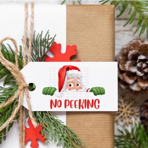 May include: A wrapped gift with a white tag featuring Santa Claus peeking over the top. The tag reads "NO PEEKING" in red letters. The gift is decorated with greenery, twine, and red snowflake accents.