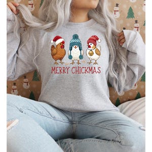 May include: A light gray sweatshirt featuring three cartoon chickens wearing festive hats. The chickens are brown, white, and brown and white. The text "MERRY CHICKMAS" is printed in red below the chickens. The sweatshirt is a casual holiday design.