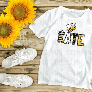 May include: White t-shirt with a bee and the name "KATE" spelled out in yellow and black letters with a bee pattern. The shirt is on a wooden surface with sunflowers.