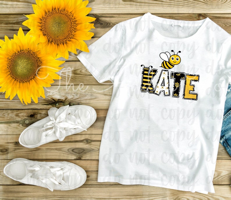 May include: White t-shirt with a bee and the name "KATE" spelled out in yellow and black letters with a bee pattern. The shirt is on a wooden surface with sunflowers.