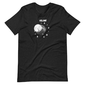 Op de afbeelding: Donkergrijs t-shirt met een afbeelding van een volumeknop en het woord "VOLUME" erboven. De knop staat op het nummer 6. De nummers 0, 8, 10 en 11 zijn ook zichtbaar.
