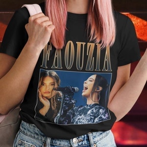 May include: Black t-shirt featuring the name "FAOUZIA" in gold lettering above a graphic of a singer. The shirt is worn with light blue jeans and a pink backpack. The person is smiling and giving a peace sign.
