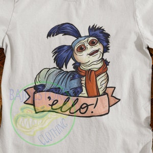 Op de afbeelding: Wit T-shirt met een cartoonillustratie van een blauwe en witte rups met een rode sjaal. De rups zegt "'ello!" in een spraakballon.