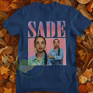May include: A navy blue t-shirt with a pink and white graphic of the singer Sade. The graphic features a portrait of Sade with the text "SADE" above her.