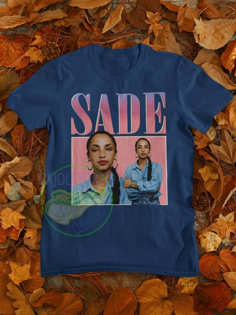 May include: A navy blue t-shirt with a pink and white graphic of the singer Sade. The graphic features a portrait of Sade with the text "SADE" above her.