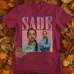 May include: A maroon t-shirt with a vintage-style graphic of Sade, a singer, with the text "SADE" in a pink and blue gradient.