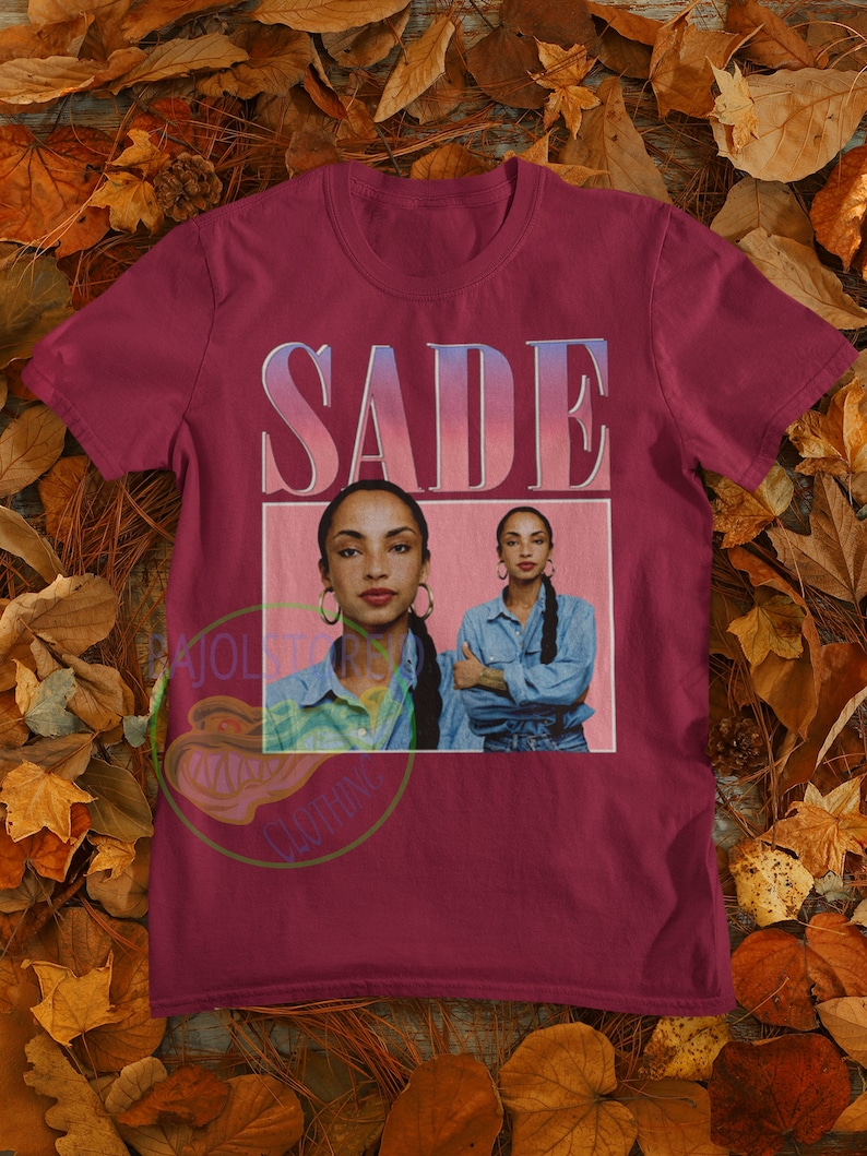 May include: A maroon t-shirt with a vintage-style graphic of Sade, a singer, with the text "SADE" in a pink and blue gradient.