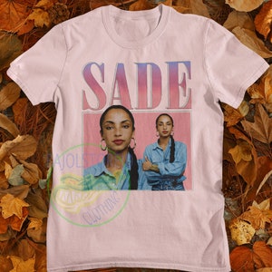 May include: Pink t-shirt with a graphic print of the singer Sade. The print features a pink background with the word "SADE" in a white outline and a photo of Sade in a blue denim shirt.