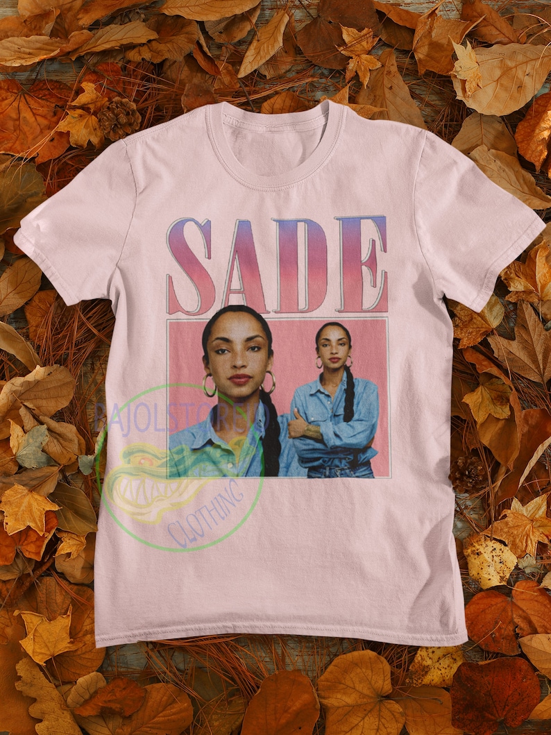May include: Pink t-shirt with a graphic print of the singer Sade. The print features a pink background with the word "SADE" in a white outline and a photo of Sade in a blue denim shirt.