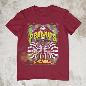 May include: A red t-shirt with a graphic of a circus tent with the words "Primus" and "Dinosaur Jr." in yellow. The graphic also includes a skull wearing a top hat and bow tie. The text below the graphic reads "August 9, 2015 - Rose Music Center - Huber Heights, OH."