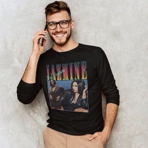 May include: A black long-sleeved shirt featuring a graphic print of the singer Jazmine Sullivan. The graphic is in a retro style with a rainbow colour scheme and the text "Jazmine" in a bold font.