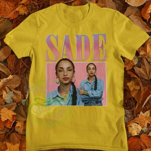 May include: Yellow t-shirt with a pink and purple graphic of the singer Sade. The graphic features a photo of Sade with the text "SADE" above her.