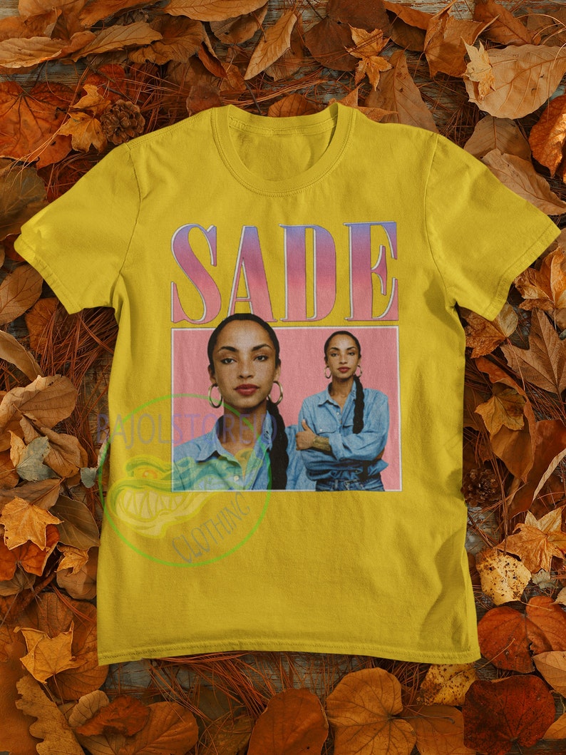 May include: Yellow t-shirt with a pink and purple graphic of the singer Sade. The graphic features a photo of Sade with the text "SADE" above her.