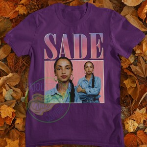 May include: Purple t-shirt with a vintage-style graphic of Sade, a singer, with the text "SADE" in pink letters.