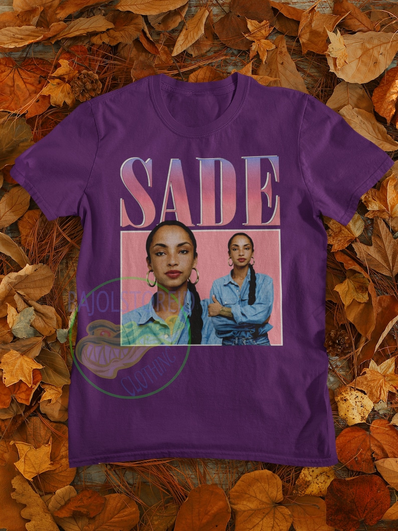 May include: Purple t-shirt with a vintage-style graphic of Sade, a singer, with the text "SADE" in pink letters.