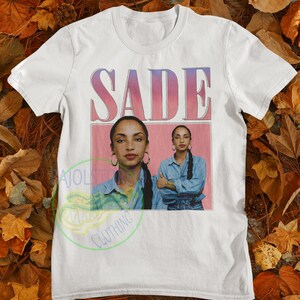 May include: White t-shirt with a pink and blue gradient graphic of the word "SADE" and a photo of Sade in a denim shirt.