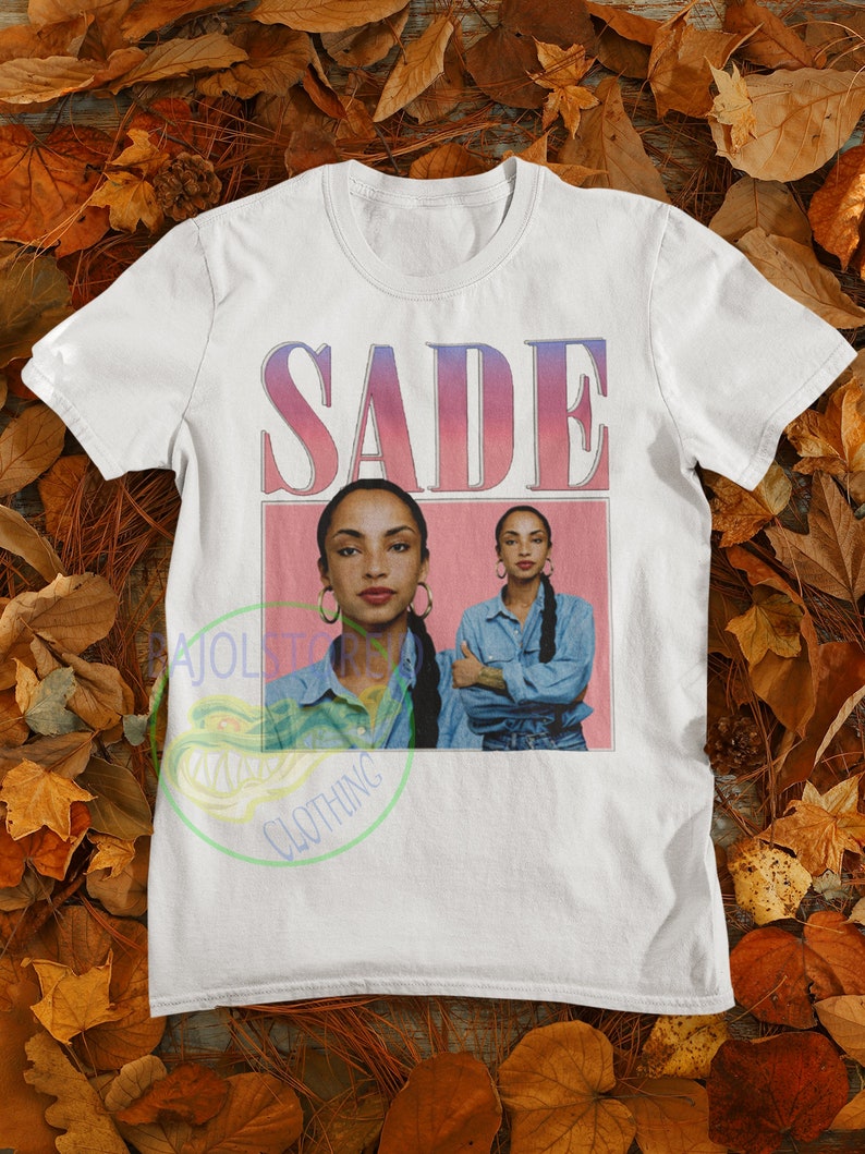 May include: White t-shirt with a pink and blue gradient graphic of the word "SADE" and a photo of Sade in a denim shirt.