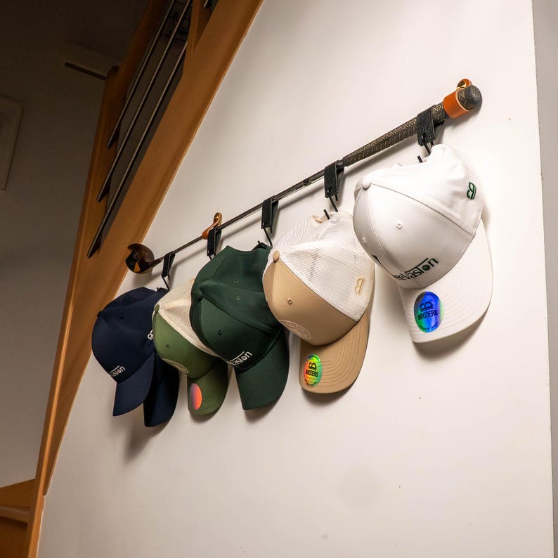 Peut inclure: Un porte-chapeaux en m&eacute;tal avec six casquettes de baseball suspendues. Les casquettes sont de diff&eacute;rentes couleurs, dont le marine, le vert et le blanc. Le porte-chapeaux est fix&eacute; &agrave; un mur blanc.