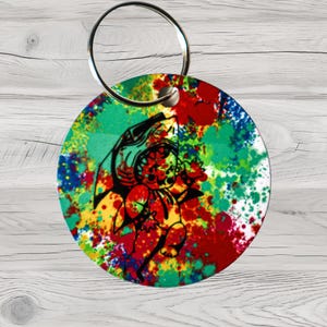 May include: Round keychain with a colorful splatter paint background. A black and white cartoon shark with a toothy grin is in the center of the keychain.