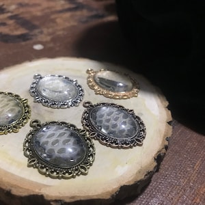 以下が含まれることがあります： 装飾的なフレームが付いた、銀と金の5つの楕円形のペンダント。各ペンダントには、白い模様が入った透明なレジンインサートが特徴です。