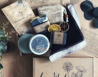 May include: A brown cardboard box with the words "Welcome to your Wild Bloom Box" printed on the top. The box is open and contains a variety of natural beauty products, including a bar of soap, a jar of body scrub, a bag of clay, a face mask, and a washcloth. The products are arranged on a bed of dried flowers and leaves.