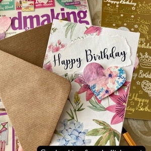 May include: A handmade card featuring a floral design and a heart-shaped embellishment. The card reads "Happy Birthday" in black script. The card is surrounded by craft supplies, including a magazine, an envelope, and a sheet of gold foil stickers.