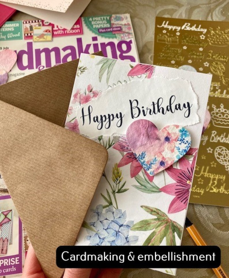 May include: A handmade card featuring a floral design and a heart-shaped embellishment. The card reads "Happy Birthday" in black script. The card is surrounded by craft supplies, including a magazine, an envelope, and a sheet of gold foil stickers.