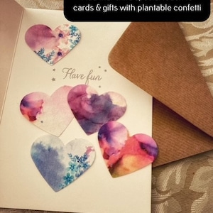 May include: A white card with the text "Have fun" and five heart-shaped confetti pieces. The confetti is decorated with watercolour-like designs in shades of pink, purple, and blue. The card is on a brown surface.