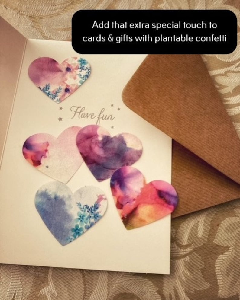 May include: A white card with the text "Have fun" and five heart-shaped confetti pieces. The confetti is decorated with watercolour-like designs in shades of pink, purple, and blue. The card is on a brown surface.
