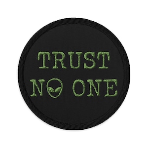 May include: A black round patch with green embroidered text that reads "TRUST NO ONE" with an alien face symbol in place of the "O" in "NO".
